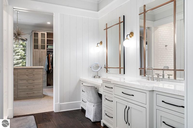 This primary bathroom features a double vanity with white cabinetry, quartz countertops, and modern black hardware. The mirrors are framed with a unique copper and black design, complemented by globe sconces. A separate vanity area with a makeup mirror and upholstered chair adds a touch of luxury, while the dark wood flooring provides a sophisticated contrast to the white walls.