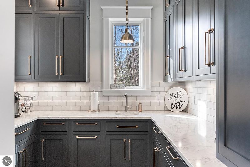 This is a well-lit kitchen featuring dark gray cabinetry with gold hardware, white subway tile backsplash, and light-colored countertops. A window above the sink provides natural light and a view of the outdoors. The kitchen has a modern and sophisticated aesthetic.