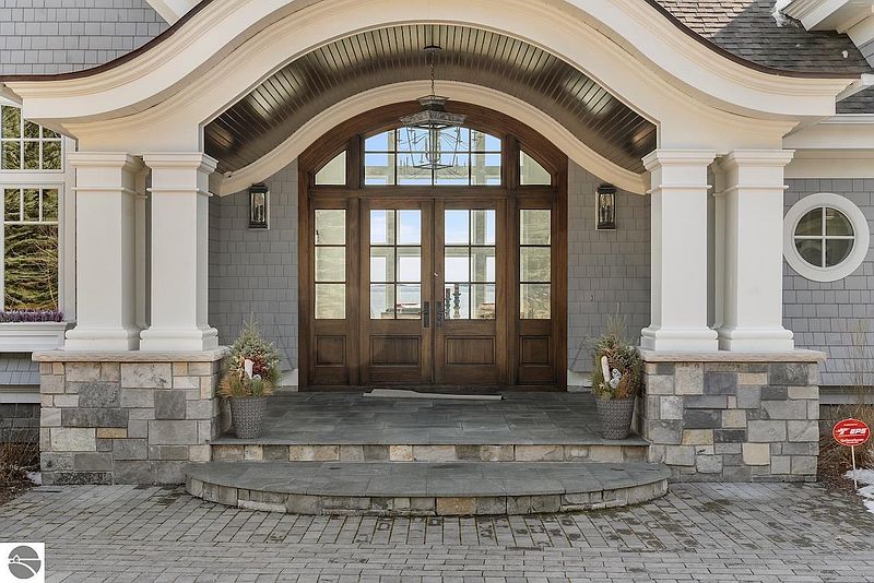 This image showcases the grand entryway of a home, featuring a large arched doorway with glass-paneled double doors and sidelights. Substantial white columns with stone bases support the arched roof, creating a welcoming and impressive facade. The entryway is accessed by stone steps, adding to the home's curb appeal.