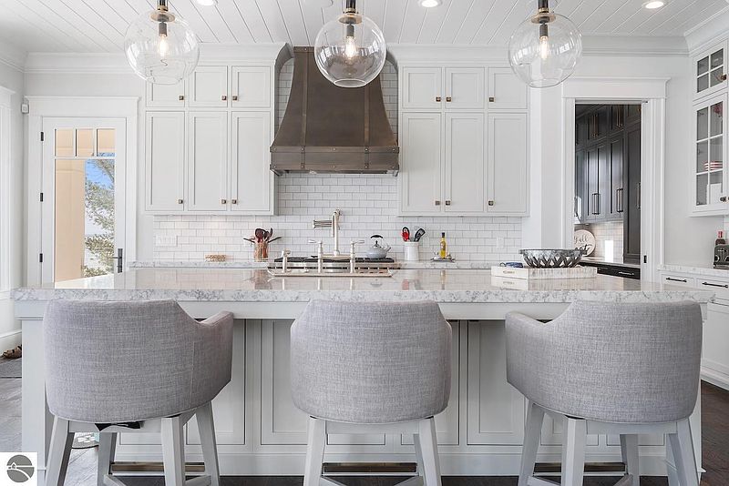 This is a bright and stylish kitchen featuring white cabinetry, a subway tile backsplash, and a large island with seating. The kitchen is illuminated by pendant lighting and includes a stainless steel range hood. The overall impression is clean, modern, and inviting.