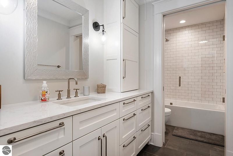 This is a well-lit bathroom featuring a white vanity with a marble countertop, complemented by brushed nickel fixtures and hardware. A decorative mirror hangs above the sink, and a glass-enclosed shower/tub is visible in the background, showcasing white subway tiles. The overall impression is clean, modern, and inviting.