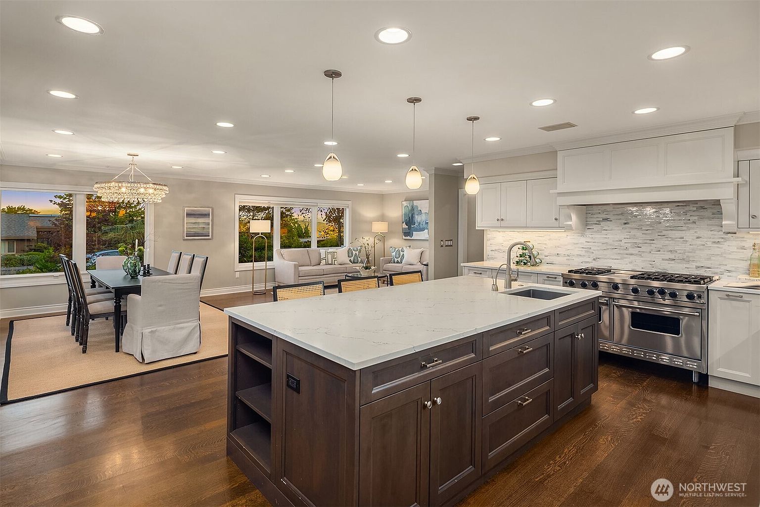 This is a wide-angle shot of a modern, open-concept kitchen that flows into a dining and living area. The kitchen features a large island with a white countertop and dark wood cabinetry, stainless steel appliances, and white upper cabinets. The dining area includes a chandelier and a view of the outdoors, while the living area has comfortable seating and natural light, creating a cohesive and inviting space.