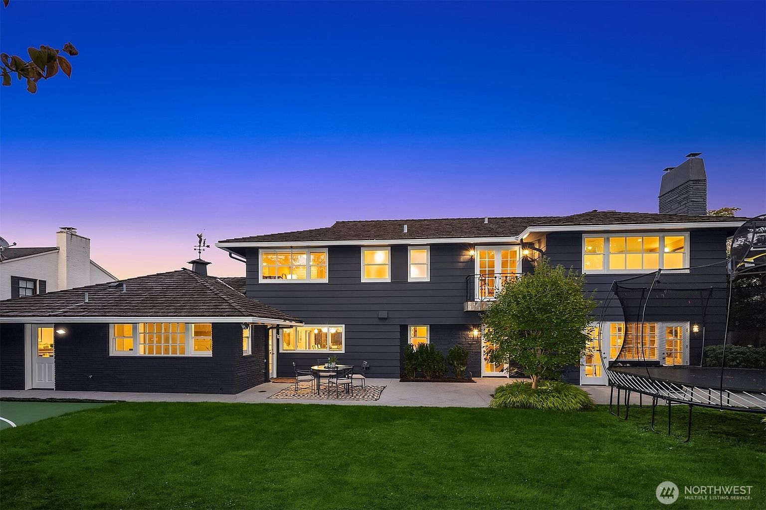 This is a rear view of a two-story house with a dark exterior and white trim, featuring a well-maintained lawn and a patio area with outdoor seating. The house has multiple windows with warm interior lighting visible, and a trampoline is set up in the yard. The image captures a serene and inviting backyard setting, perfect for relaxation and entertainment.