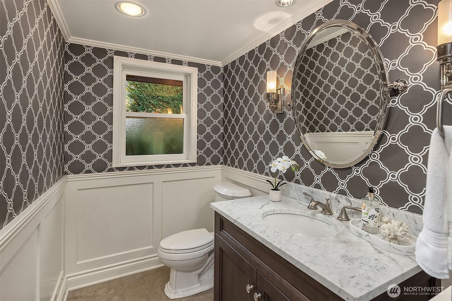 This is a well-appointed guest bathroom featuring a dark wood vanity with a marble countertop and an oval sink. The walls are decorated with a gray and white patterned wallpaper above white wainscoting. A silver-framed oval mirror hangs above the sink, and a window provides natural light, creating a sophisticated and inviting space.