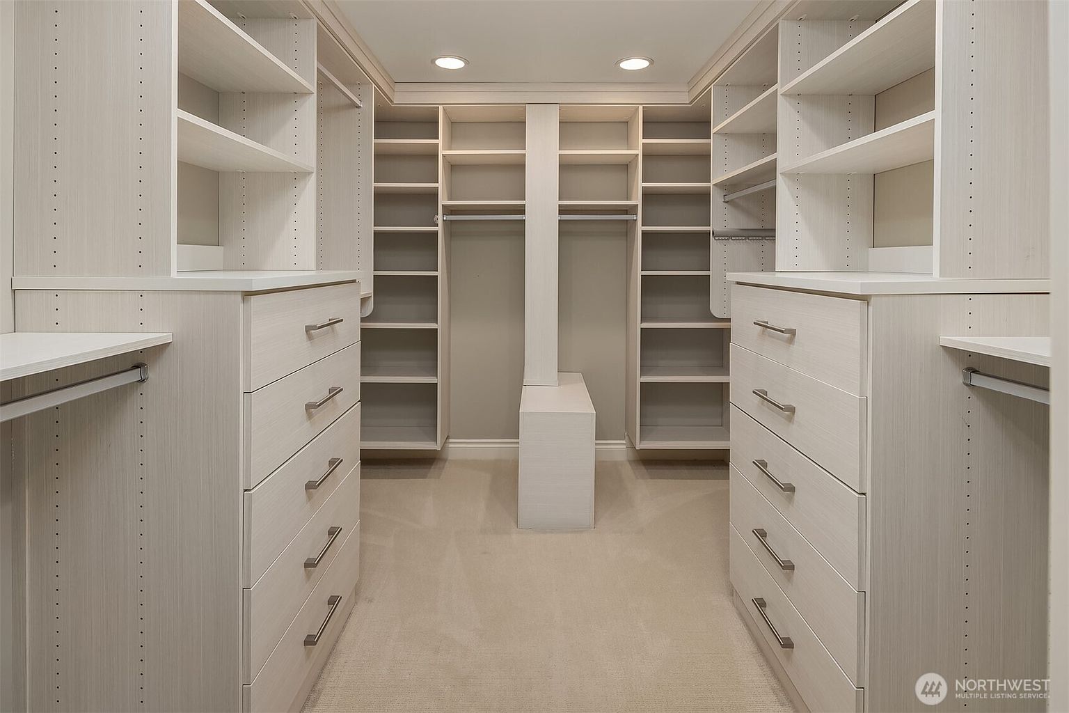 This is a well-organized walk-in closet featuring custom shelving and drawer units in a light, neutral color. The closet is spacious, with ample storage for clothing and accessories. The design is clean and modern, creating a luxurious and functional space.