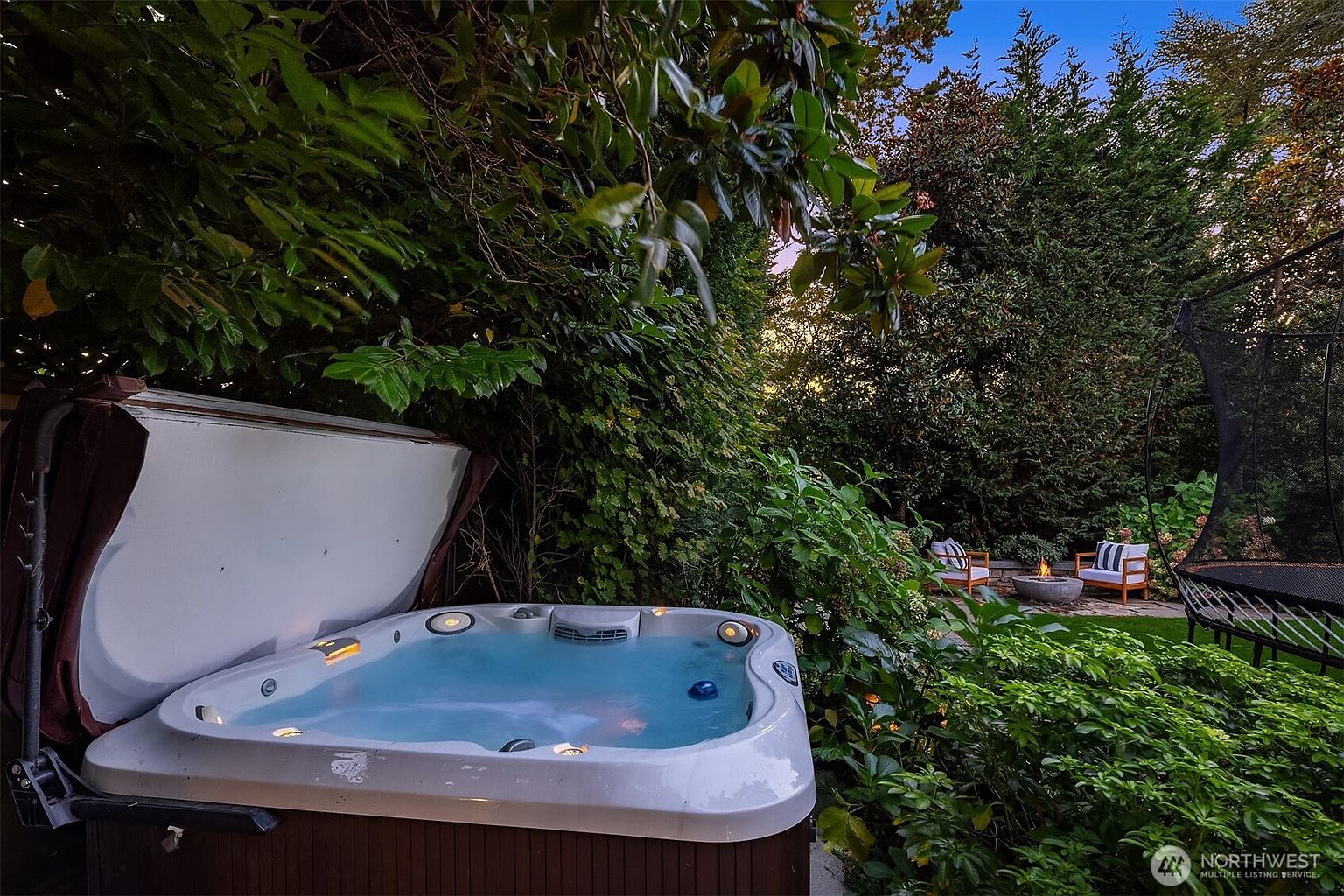 This image showcases a beautifully landscaped backyard featuring a hot tub with its cover partially open, inviting relaxation. In the background, a cozy fire pit area with chairs and a trampoline suggest entertainment and leisure. Lush greenery surrounds the space, creating a private and serene outdoor oasis.