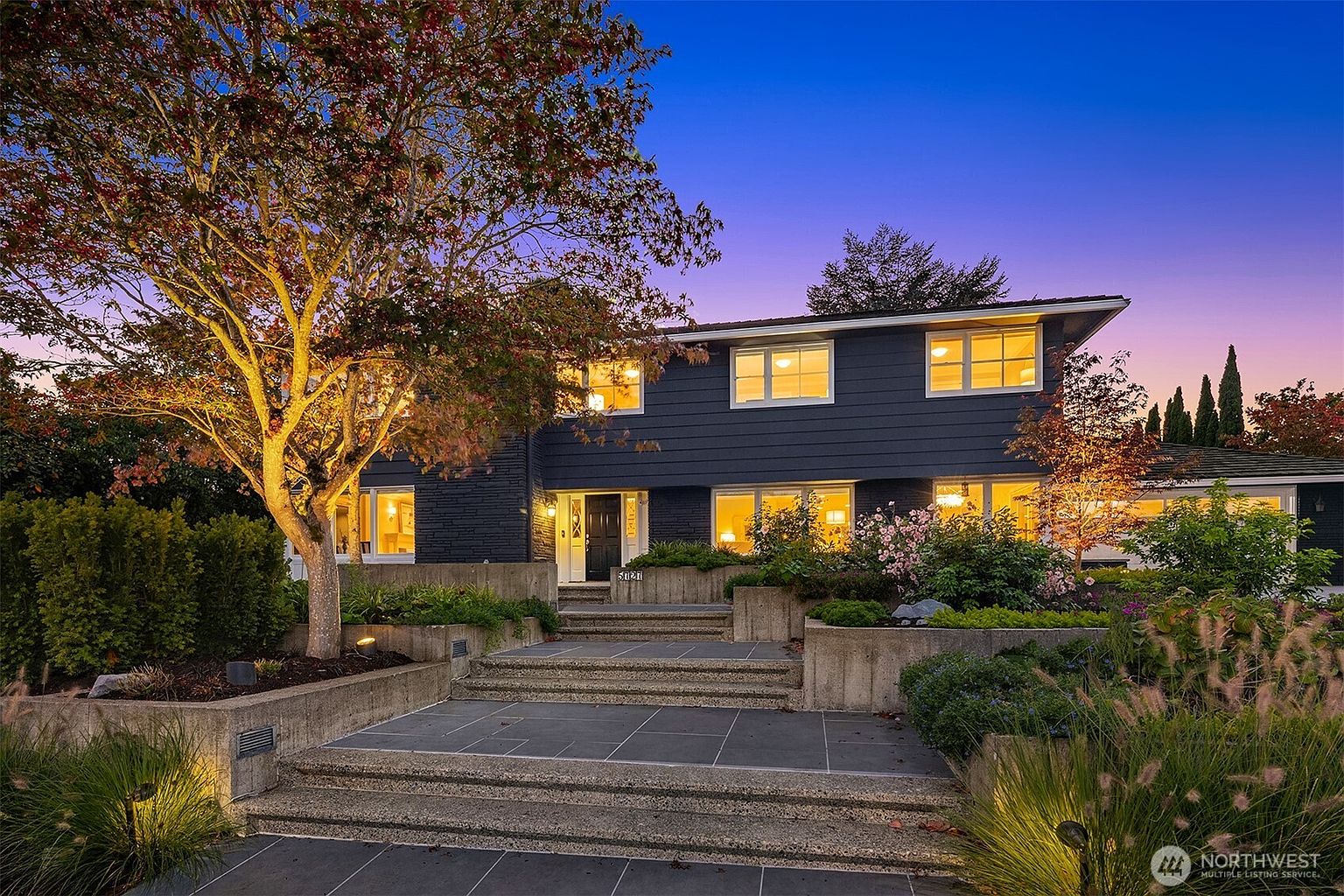 This is a captivating front view of a two-story home at twilight, showcasing a well-manicured landscape and inviting entryway. The house features a dark exterior with bright, illuminated windows, creating a warm and welcoming ambiance. A mature tree with illuminated branches adds character to the property, while the tiered stone steps lead to the front door, enhancing the curb appeal.