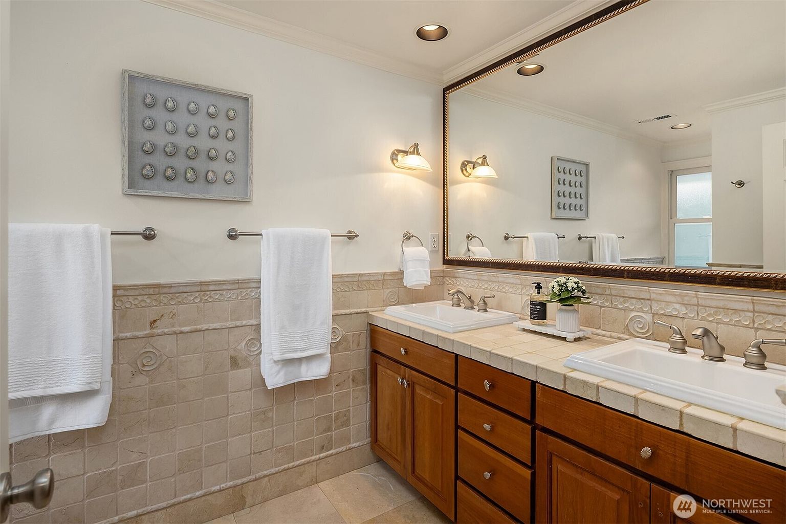 This is a well-lit bathroom featuring a double vanity with a light-colored tile countertop and wooden cabinets. The walls are adorned with decorative tiles and a framed art piece hangs above a towel rack. A large mirror reflects the scene, enhancing the sense of space.