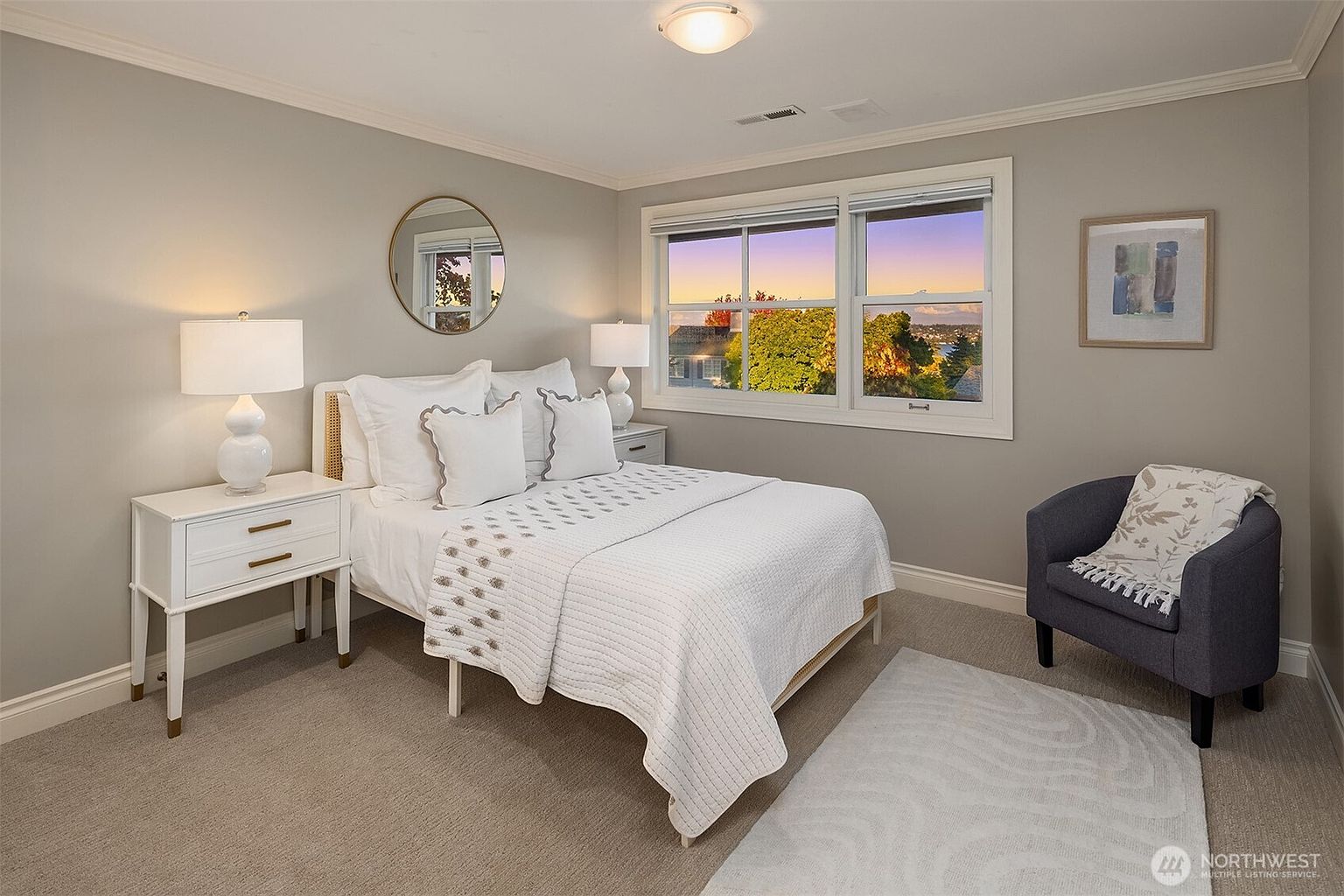 This is a well-lit primary bedroom featuring a bed with white linens and decorative pillows, flanked by a white nightstand with a lamp. A round mirror hangs above the bed, and a window provides natural light and a view of the outdoors. A gray armchair with a patterned throw sits in the corner, adding a cozy touch to the room.