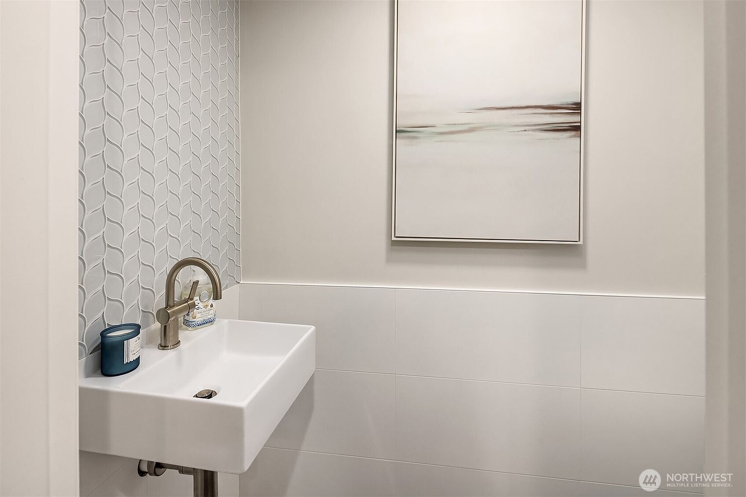 This is a well-lit guest bathroom featuring a modern, wall-mounted square sink with a brushed nickel faucet. The wall behind the sink is adorned with a decorative, light blue mosaic tile pattern, while the rest of the lower wall is covered in white horizontal tiles. An abstract art piece hangs above the tiled wall, adding a touch of sophistication to the space.