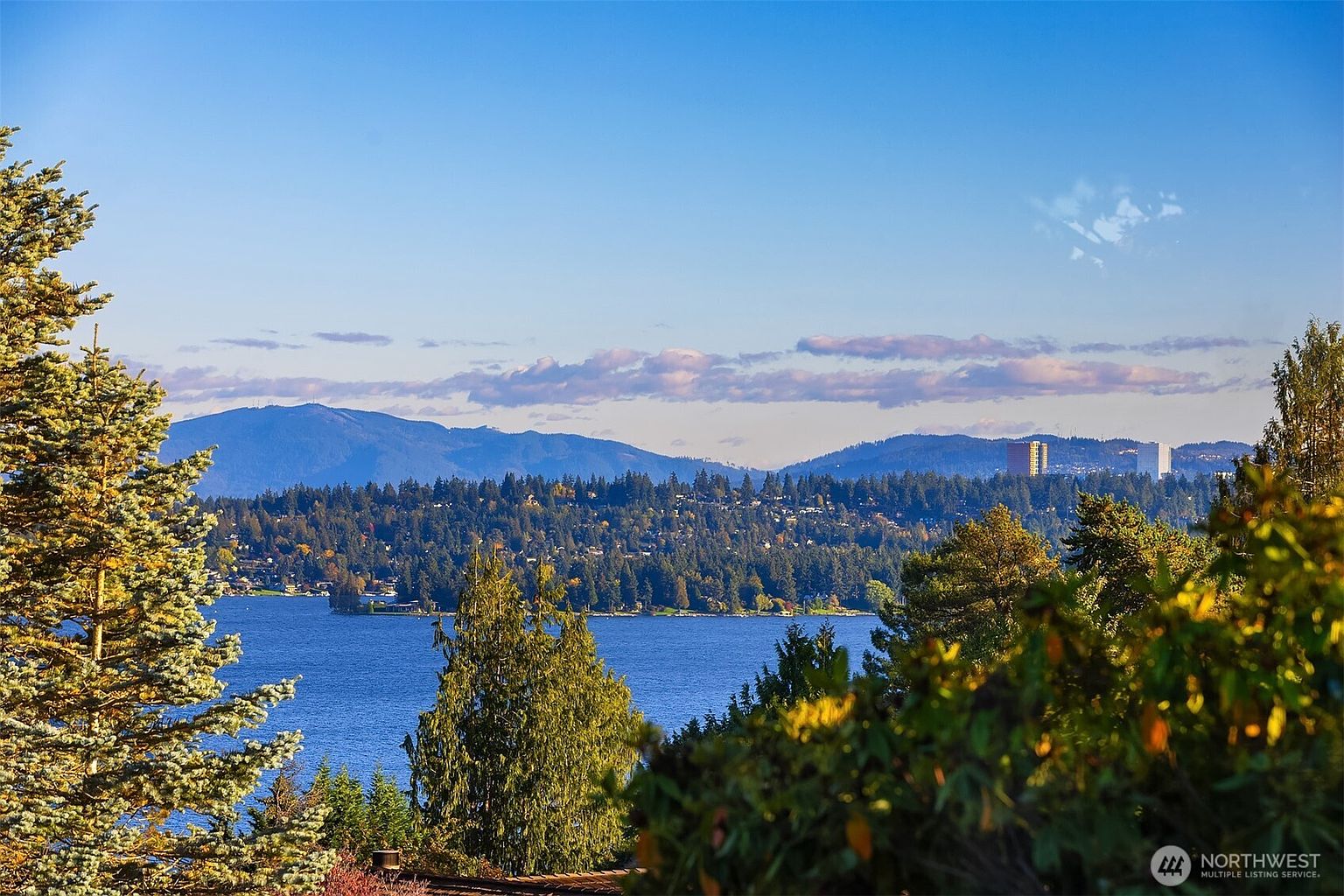 This aerial view showcases a stunning landscape featuring a serene lake surrounded by lush greenery and distant mountains under a clear blue sky with scattered clouds. The composition highlights the natural beauty and tranquility of the location, suggesting a peaceful and desirable setting for a property. A building is visible in the distance.
