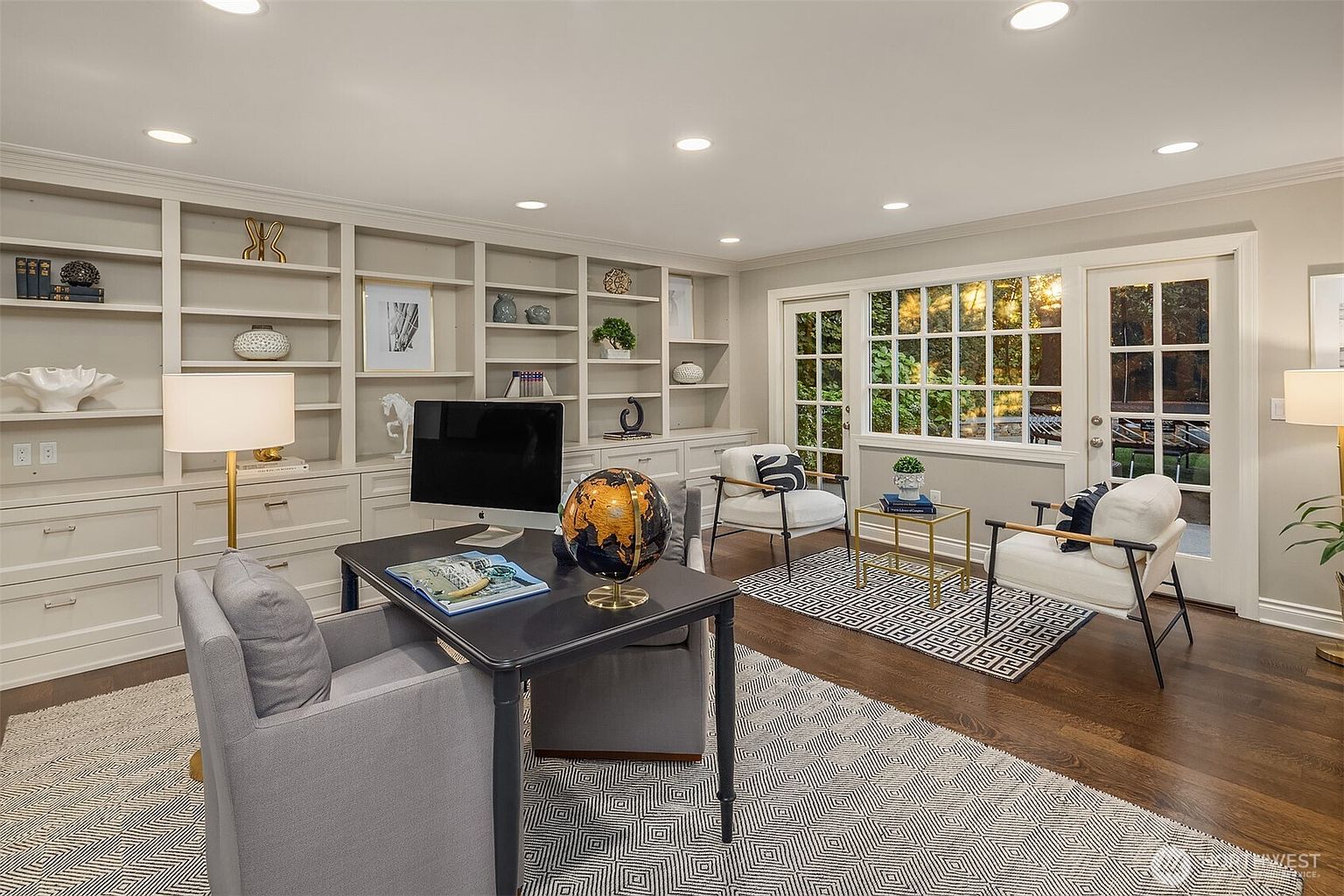 This is an interior shot of a well-appointed home office. The room features custom built-in shelving, a dark-colored desk with a globe and computer, and a comfortable gray chair. Two additional armchairs are positioned near a window and door leading to the outside, with a patterned rug tying the space together, creating a sophisticated and functional workspace.