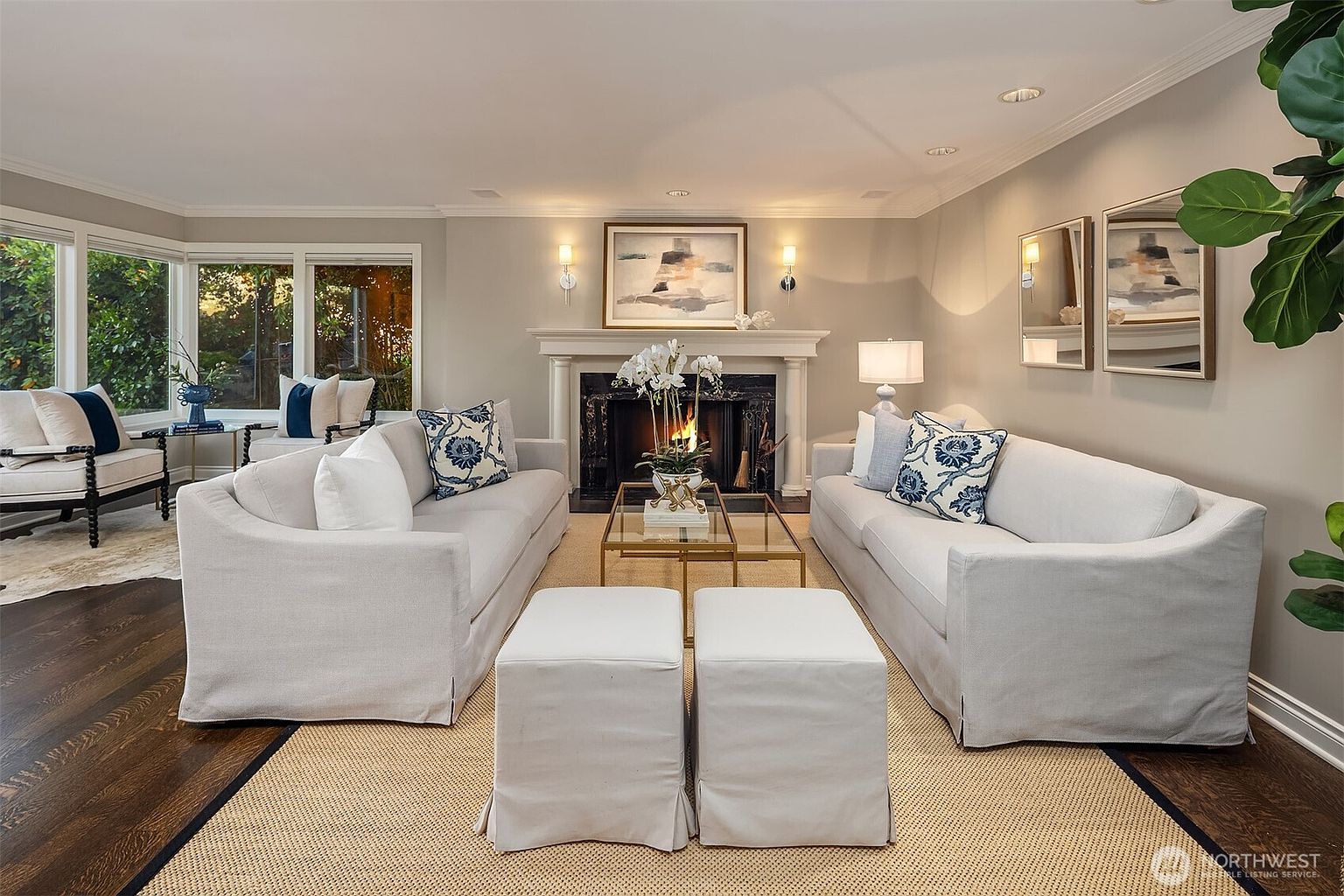 This is a well-staged living room featuring two light gray sofas facing each other, separated by a glass-topped coffee table and two white ottomans. A fireplace with a white mantel is centered on one wall, flanked by sconces and artwork. The room is bright and inviting, with hardwood floors and a neutral color palette, creating a sophisticated and comfortable atmosphere.