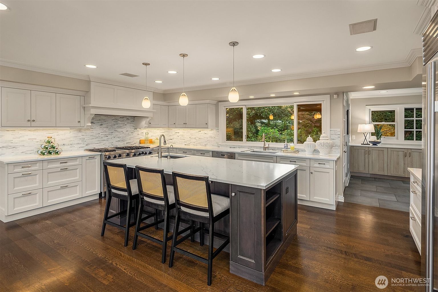 This is a bright and spacious kitchen featuring white cabinetry, a large center island with bar seating, and stainless steel appliances. The kitchen has a modern design with a light color palette, hardwood floors, and pendant lighting above the island. A large window over the sink provides natural light and a view of the outdoors.