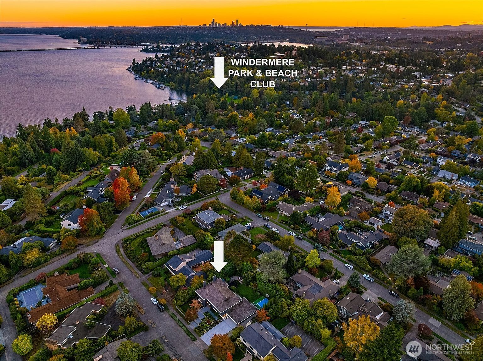 This is an aerial view of a residential neighborhood with lush greenery and a lake in the background. The houses are well-maintained with varying architectural styles, and the streets are lined with mature trees displaying autumn colors. The overall impression is one of a peaceful and upscale community.