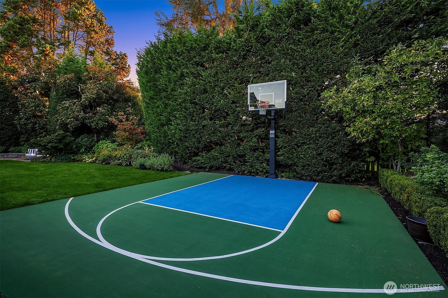 This image showcases a well-maintained backyard featuring a basketball court with a blue key and green perimeter. The court is surrounded by lush greenery, including tall hedges and trees, providing privacy and a serene atmosphere. A basketball rests on the court, suggesting recreational use.