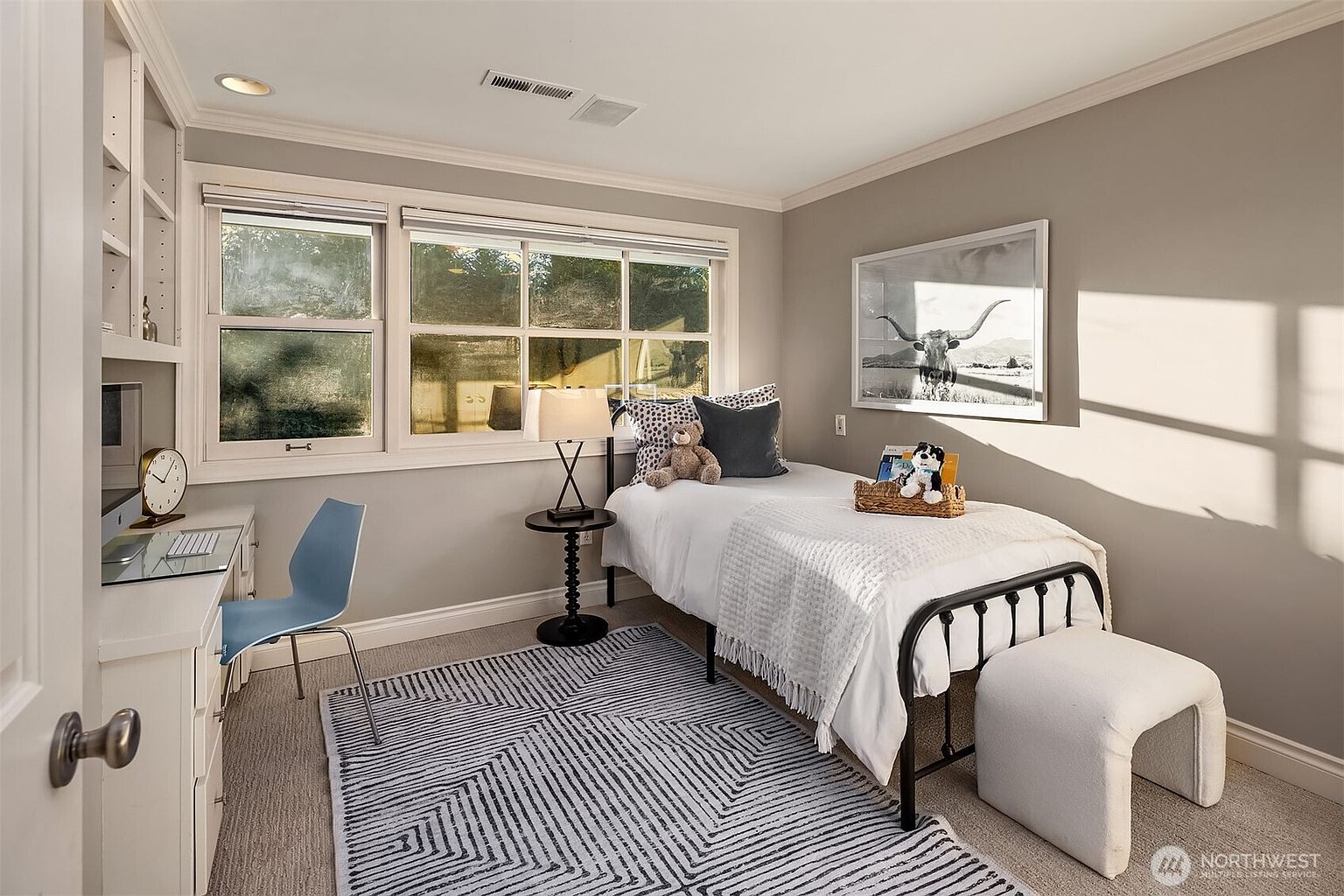 This is a cozy guest bedroom featuring a single bed with white linens and decorative pillows. A small desk area with a blue chair is positioned near the window, providing a functional workspace. The room is decorated with a black and white rug and a framed picture, creating a comfortable and inviting atmosphere.