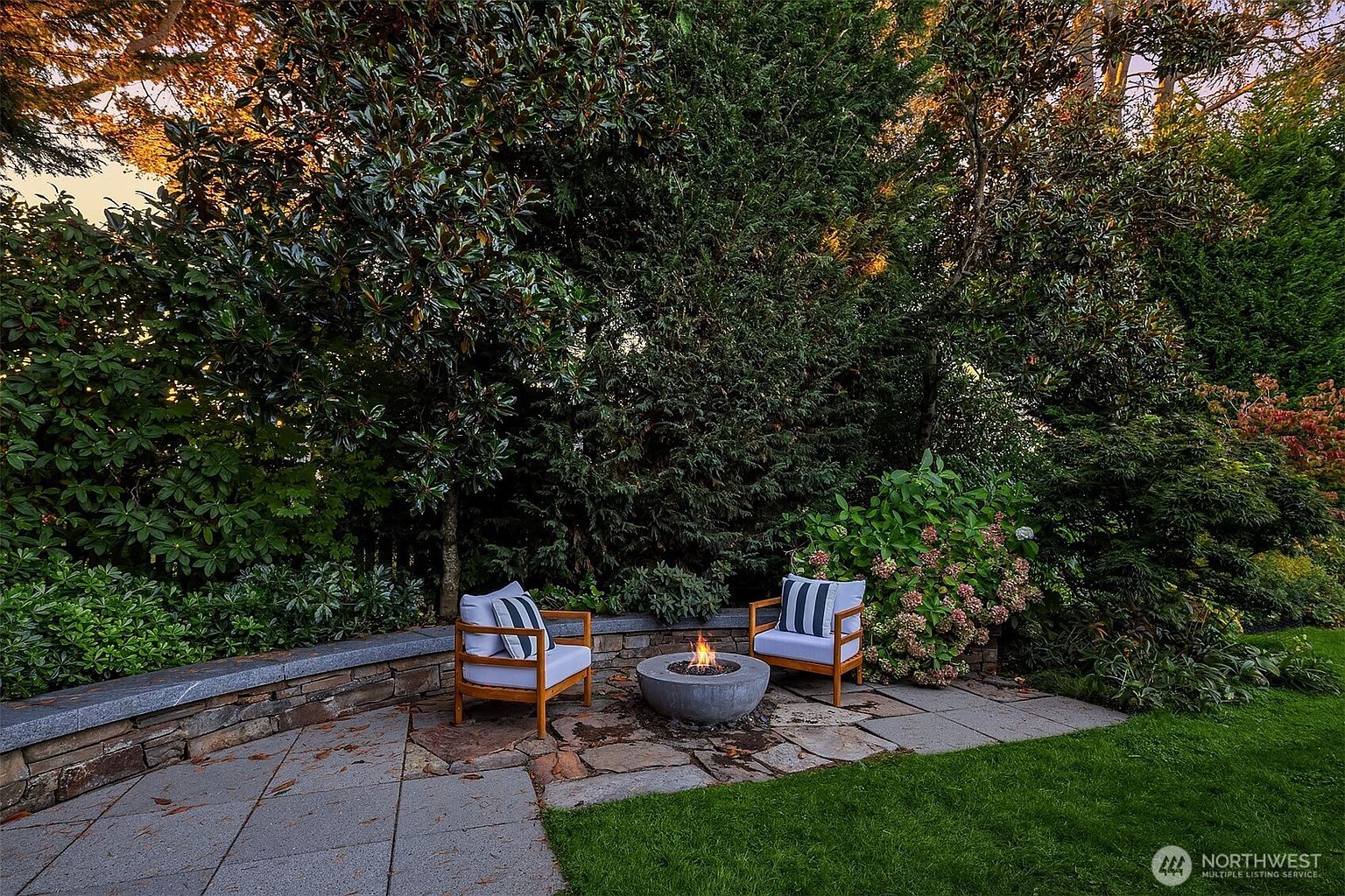 This image showcases a cozy outdoor patio area, perfect for relaxation and entertaining. Two wooden chairs with striped cushions flank a modern concrete fire pit, creating a focal point. The patio is surrounded by lush greenery, including trees, bushes, and flowers, providing privacy and a serene atmosphere. The stone patio is adjacent to a well-maintained lawn, enhancing the overall appeal of the outdoor space.