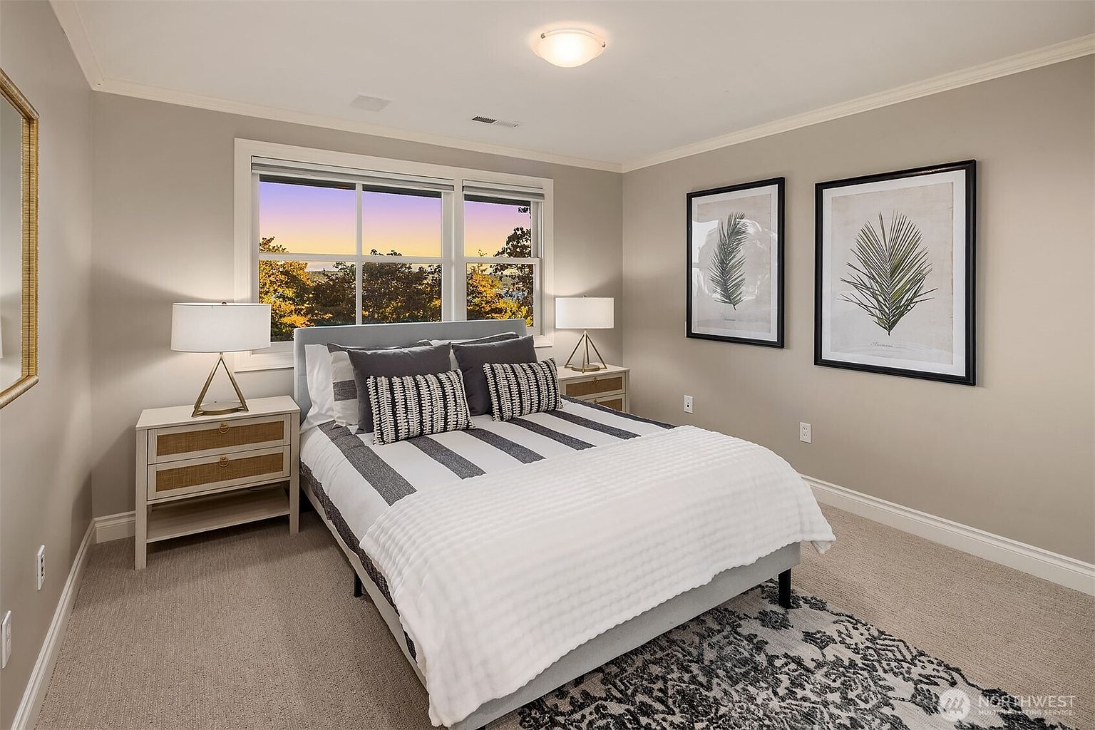 This is a cozy bedroom featuring a bed with striped bedding and decorative pillows, flanked by matching nightstands with lamps. Two framed botanical prints adorn the wall, adding a touch of nature to the space. The room is well-lit and has a neutral color palette, creating a serene and inviting atmosphere.