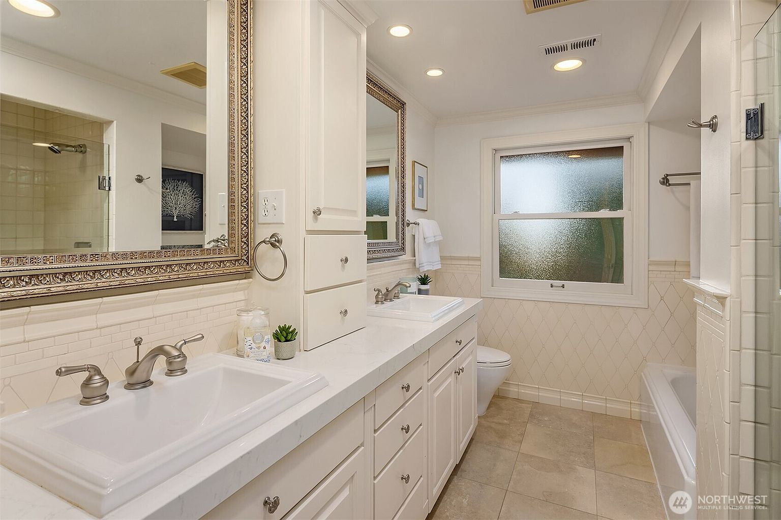 This is a well-lit primary bathroom featuring a double vanity with white countertops and cabinetry. The mirrors are framed with ornate detailing, and the walls are adorned with a patterned tile design. A window provides natural light, and the overall impression is clean and classic.