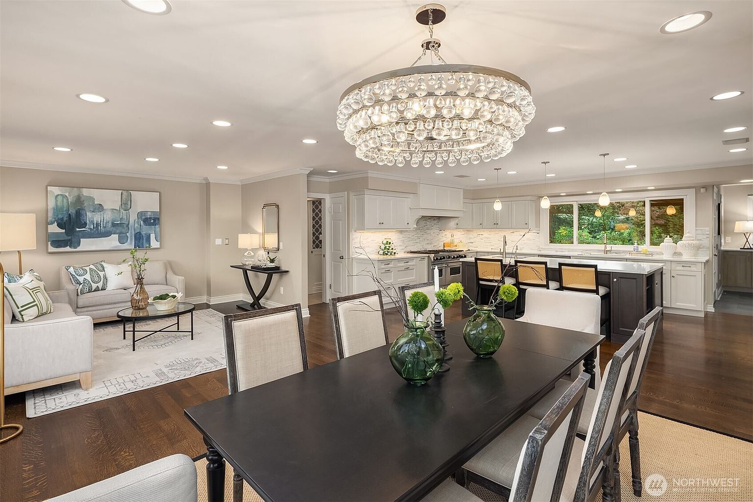 This interior shot showcases an open-concept dining area seamlessly connected to the living room and kitchen. A large, ornate chandelier hangs above a dark wood dining table, surrounded by upholstered chairs. The space is well-lit, highlighting the hardwood floors and the neutral color palette, creating an inviting and sophisticated atmosphere.