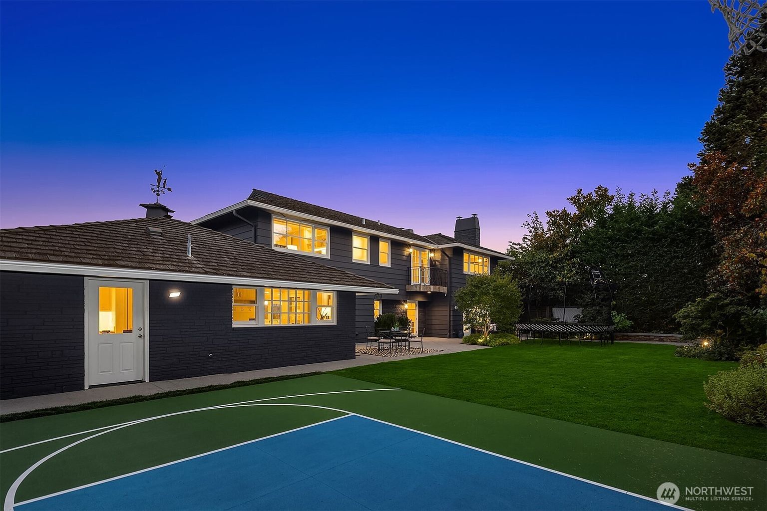 This image showcases the rear exterior of a two-story home at dusk, featuring a dark gray brick facade, a brown shingle roof, and multiple illuminated windows. A basketball court is visible in the foreground, leading to a well-manicured lawn and patio area with outdoor seating. The property is surrounded by mature trees and landscaping, creating a private and inviting backyard space.