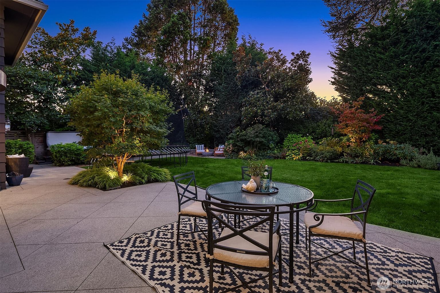 This image showcases a beautifully landscaped backyard at dusk, featuring a well-manicured lawn, mature trees, and a patio area. A round table with chairs sits on a patterned rug, creating an inviting outdoor dining space. The scene is peaceful and serene, highlighting the property's outdoor living potential.