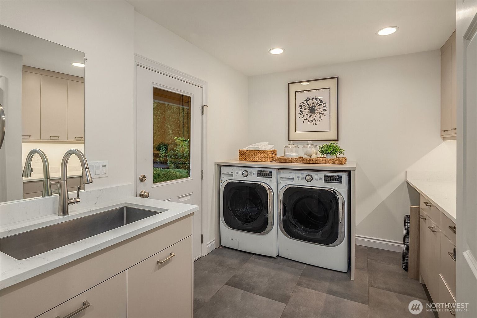 This is a well-organized laundry room featuring a stainless steel sink with a modern faucet, light-colored cabinetry, and a pair of front-loading washing machines. A countertop sits above the machines, providing space for laundry supplies and decorative items. The room is brightly lit with recessed lighting and natural light coming through a glass-paneled door, creating a clean and functional space.