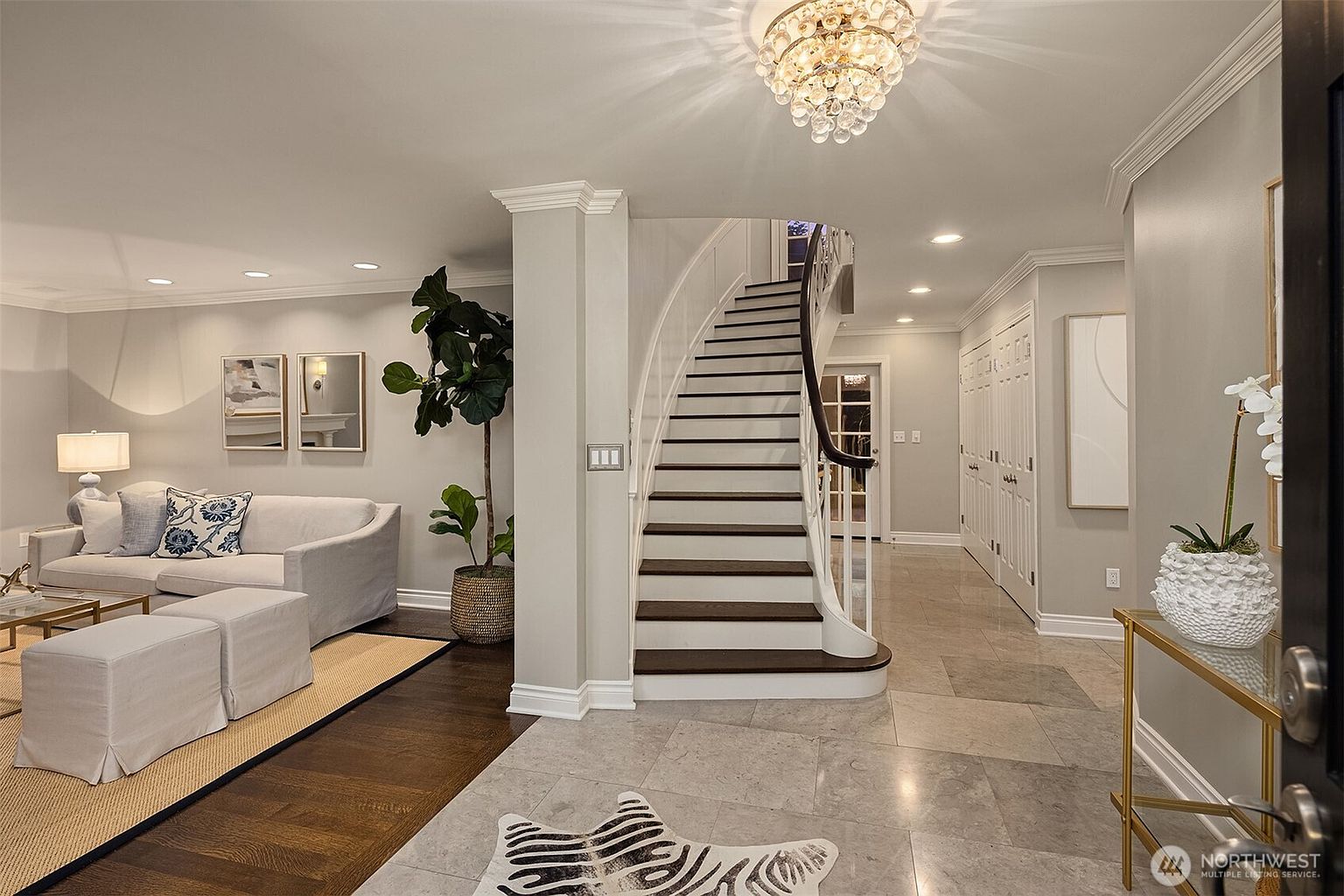 This interior shot showcases an elegant hallway and staircase. The staircase features dark wood treads and white risers, complemented by a curved white banister. The hallway is adorned with neutral-toned walls, recessed lighting, and a decorative chandelier, creating a bright and inviting atmosphere.