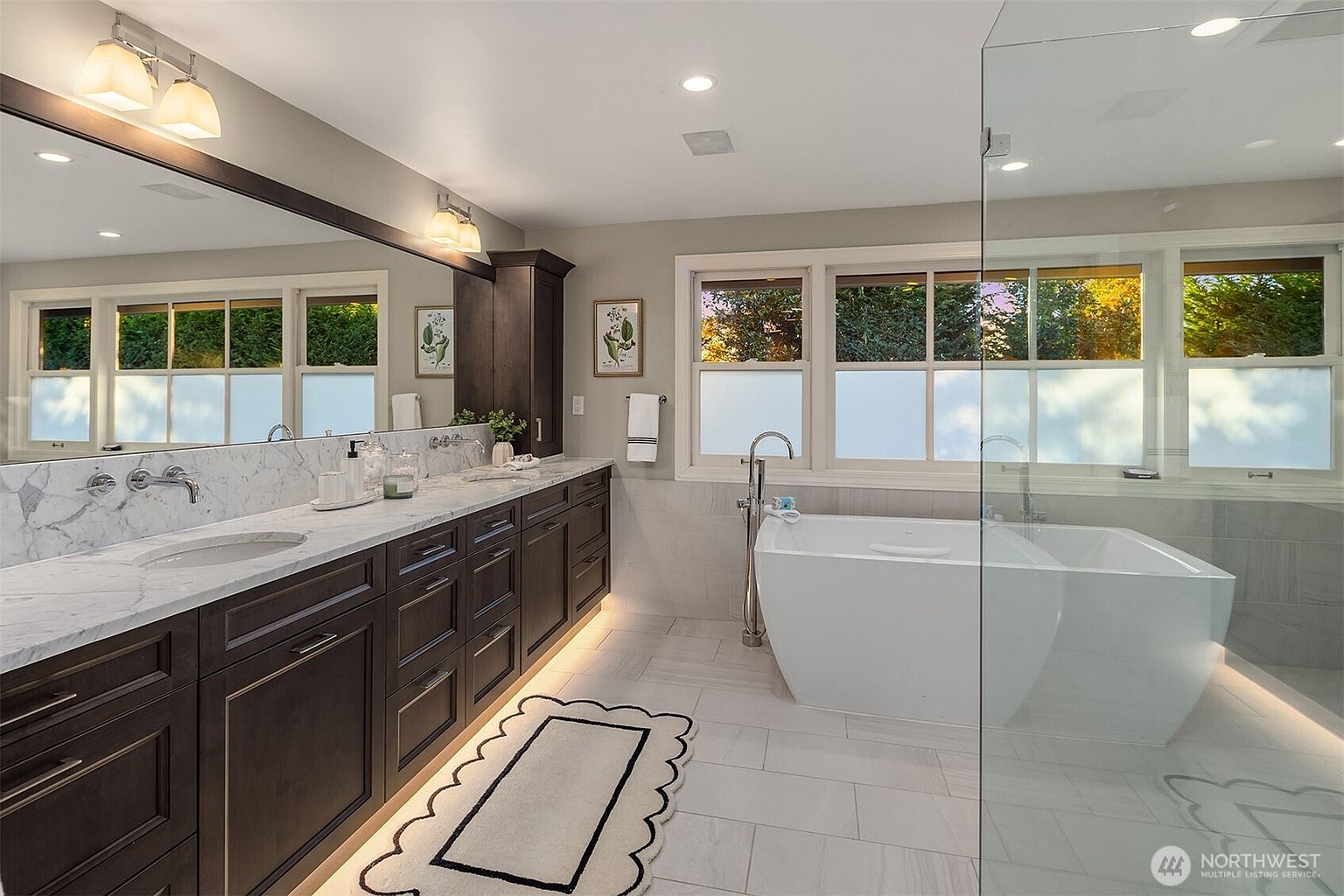 This is a well-lit primary bathroom featuring a double vanity with a marble countertop and dark wood cabinetry. A freestanding bathtub sits beneath a window with privacy film, and a glass-enclosed shower is adjacent to the tub. The flooring is light-colored tile, and a decorative rug adds a touch of elegance.