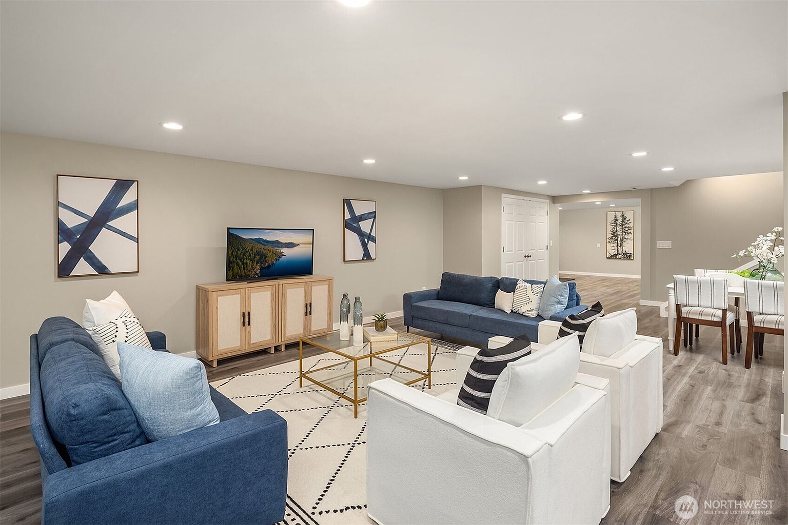This is a well-staged living room featuring two blue sofas, two white armchairs, and a gold-framed coffee table on a patterned rug. A wooden cabinet supports a television, and artwork adorns the walls. The room has a modern and inviting feel, with neutral wall colors and recessed lighting.