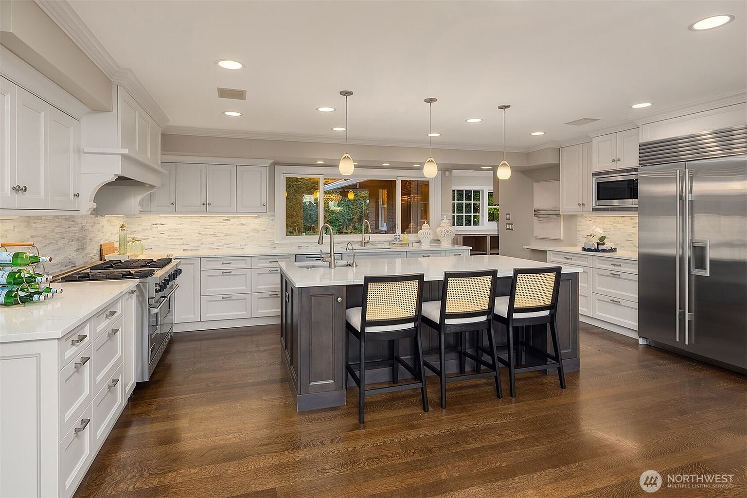 This is a bright and spacious kitchen featuring white and gray cabinetry, stainless steel appliances, and a large center island with bar seating. The kitchen is well-lit with recessed lighting and pendant lights, and it has hardwood floors. The overall impression is one of modern elegance and functionality.