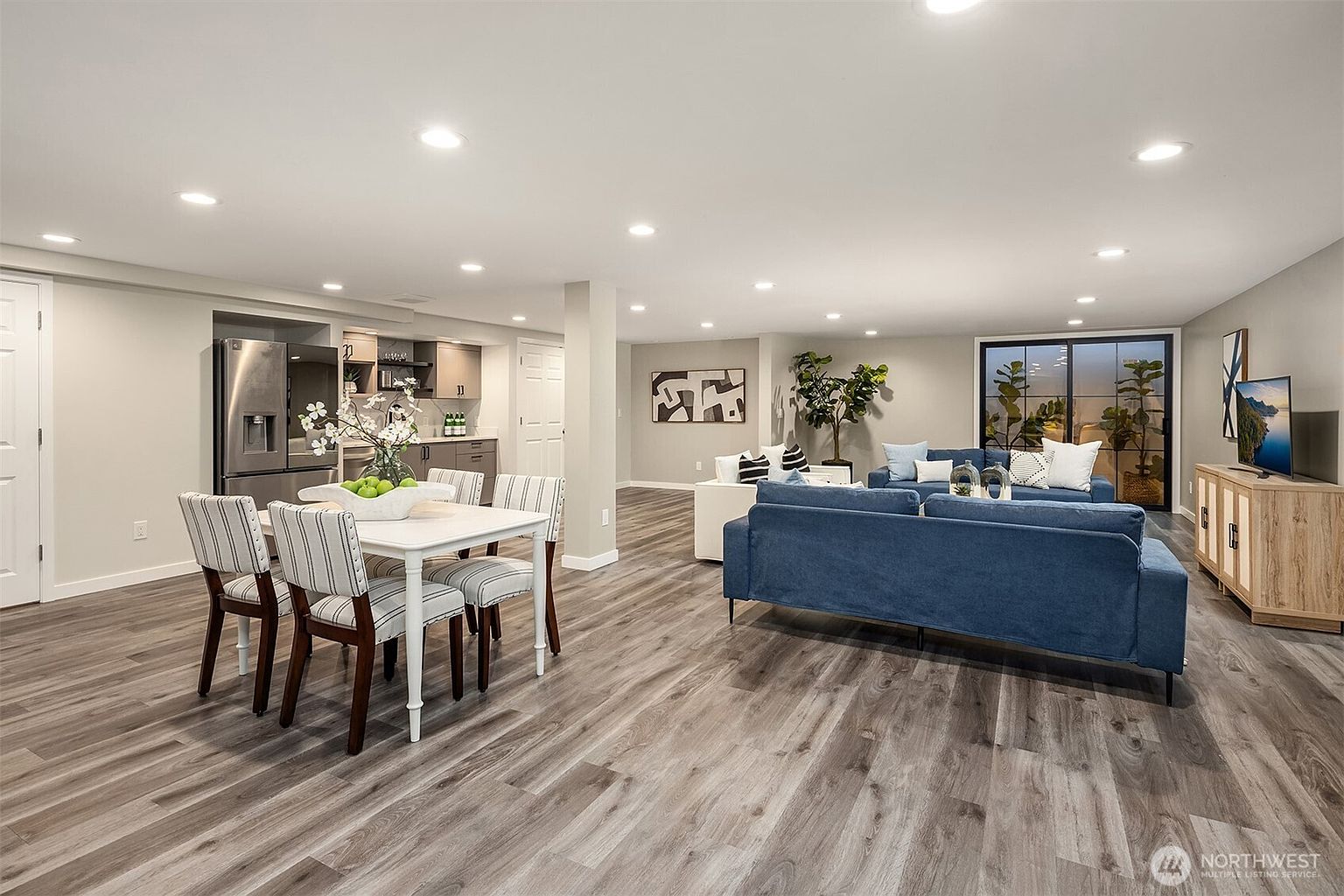 This is a spacious, open-concept living area featuring a dining space and a comfortable lounge. The room is well-lit with recessed lighting and natural light from a sliding glass door. The flooring is a modern wood-look plank, and the decor is contemporary with a blue sofa, white accent chairs, and a light wood media console.