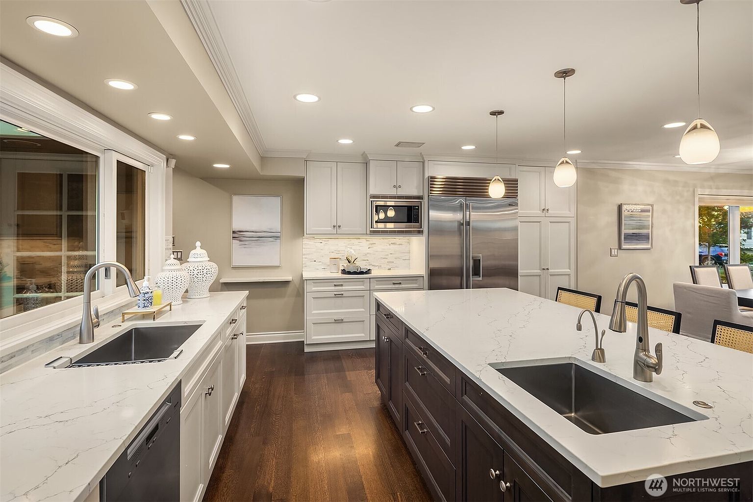 This is a well-lit kitchen featuring a large island with a sink and dark wood cabinetry, complemented by white countertops and stainless steel appliances. The kitchen also includes white cabinets along the walls and a window overlooking the exterior. The overall impression is one of modern elegance and functionality.
