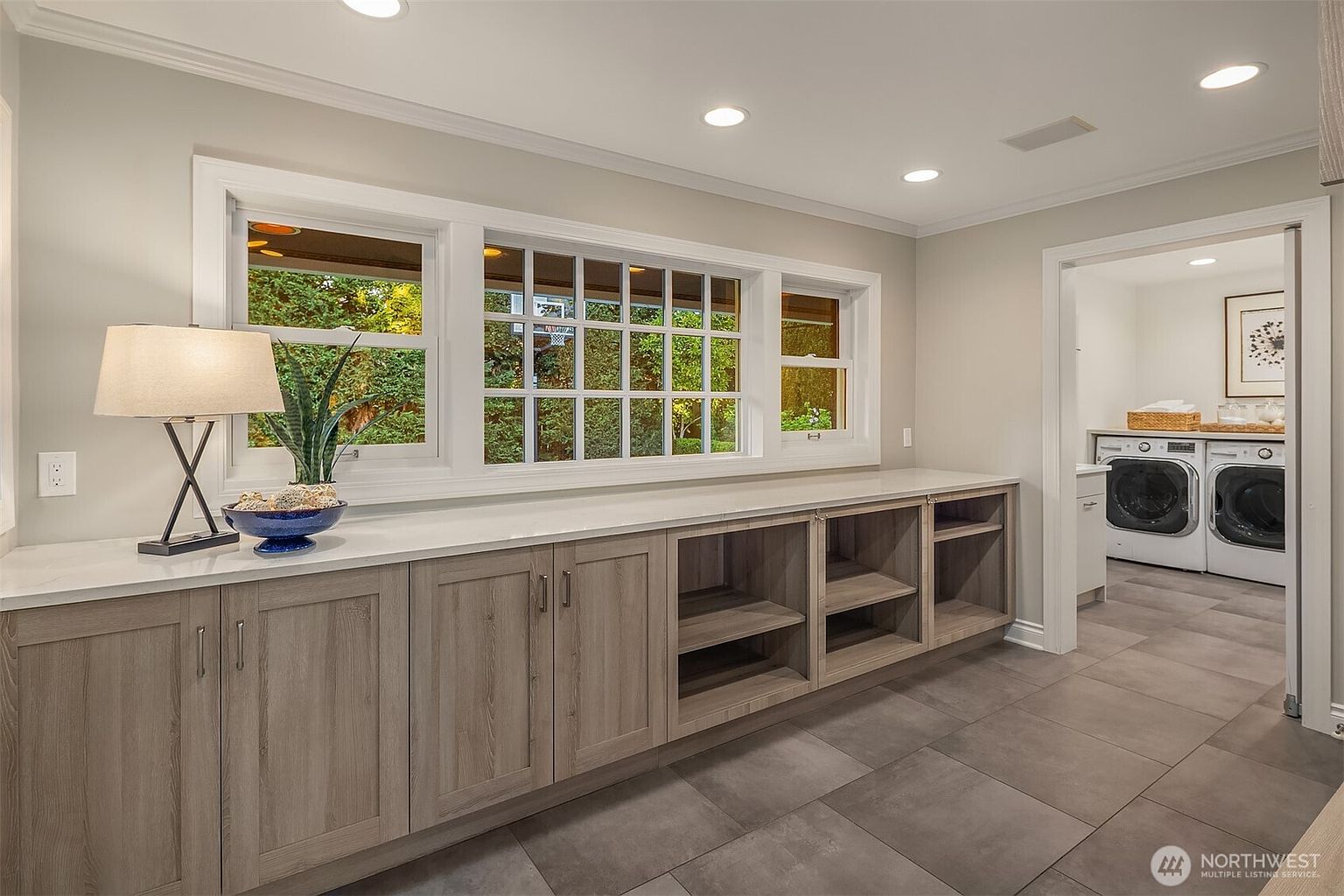 This is a well-lit laundry room featuring gray cabinetry with a white countertop, providing ample storage and workspace. A large window offers natural light and a view of the outdoors. The room leads into a separate laundry area with a washer and dryer, creating a functional and organized space.