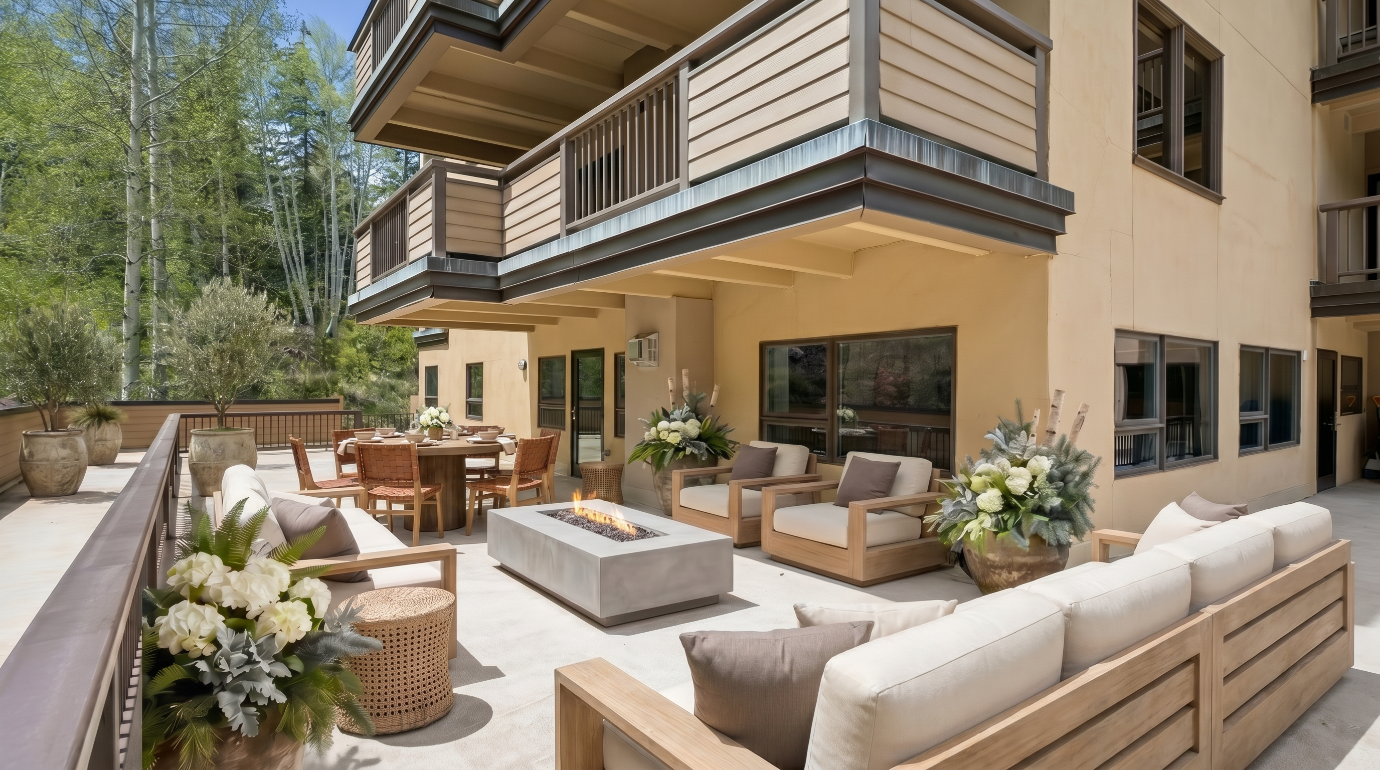 This spacious, luxury outdoor terrace features a large sectional sofa, a modern concrete fire pit, and a dining area, making it ideal for entertainment. The area is elegantly decorated with potted greenery and white floral arrangements, set against a multi-story building with a warm, neutral exterior finish. With its well-manicured ambiance and serene forest-adjacent setting, the balcony offers a sophisticated retreat for outdoor living.