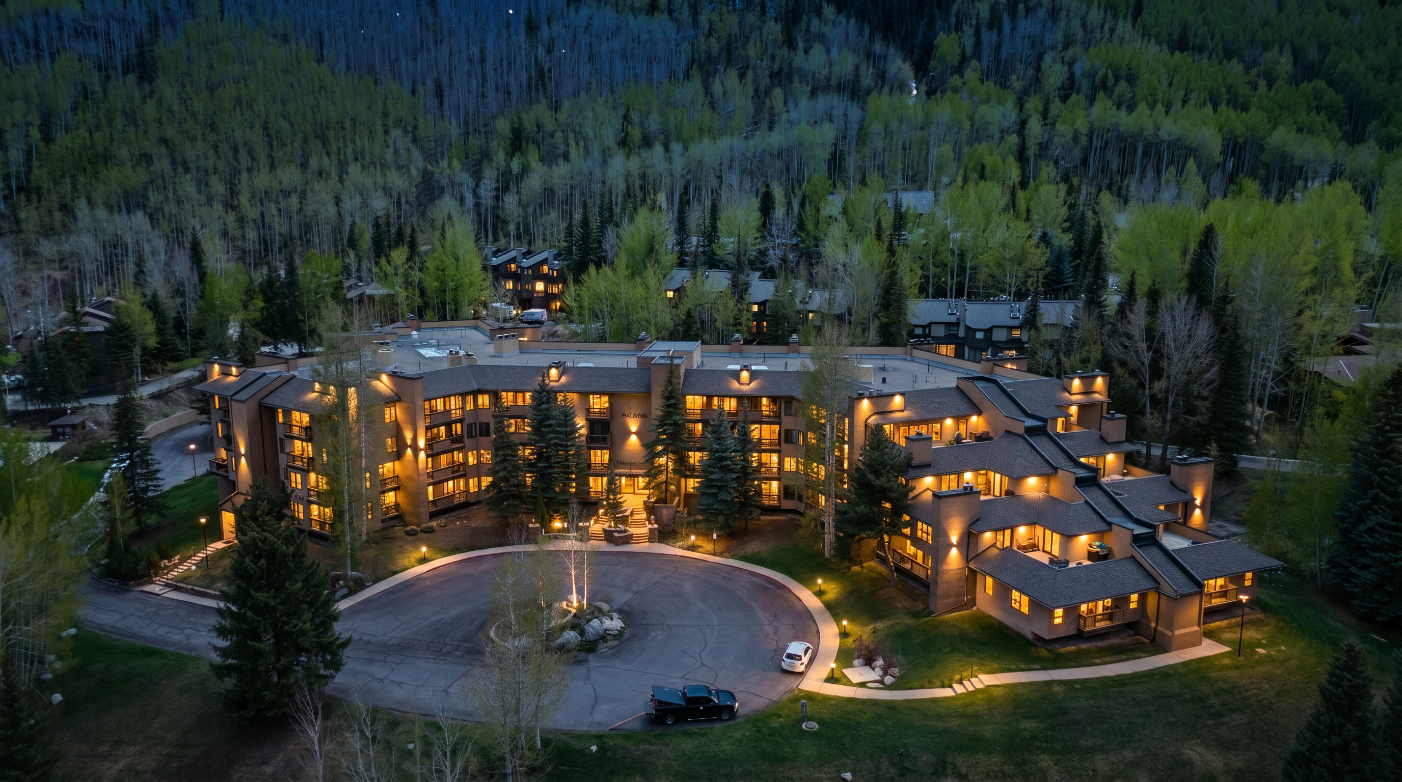 This high-angle aerial shot captures the Fall Ridge residential complex nestled within a lush, forested mountain landscape. The multi-story, earth-toned building features a sprawling architectural design with a circular driveway in the foreground and dense aspen and evergreen trees surrounding the property. The perspective provides a comprehensive view of the building's layout, roof structure, and its integration into the natural environment.