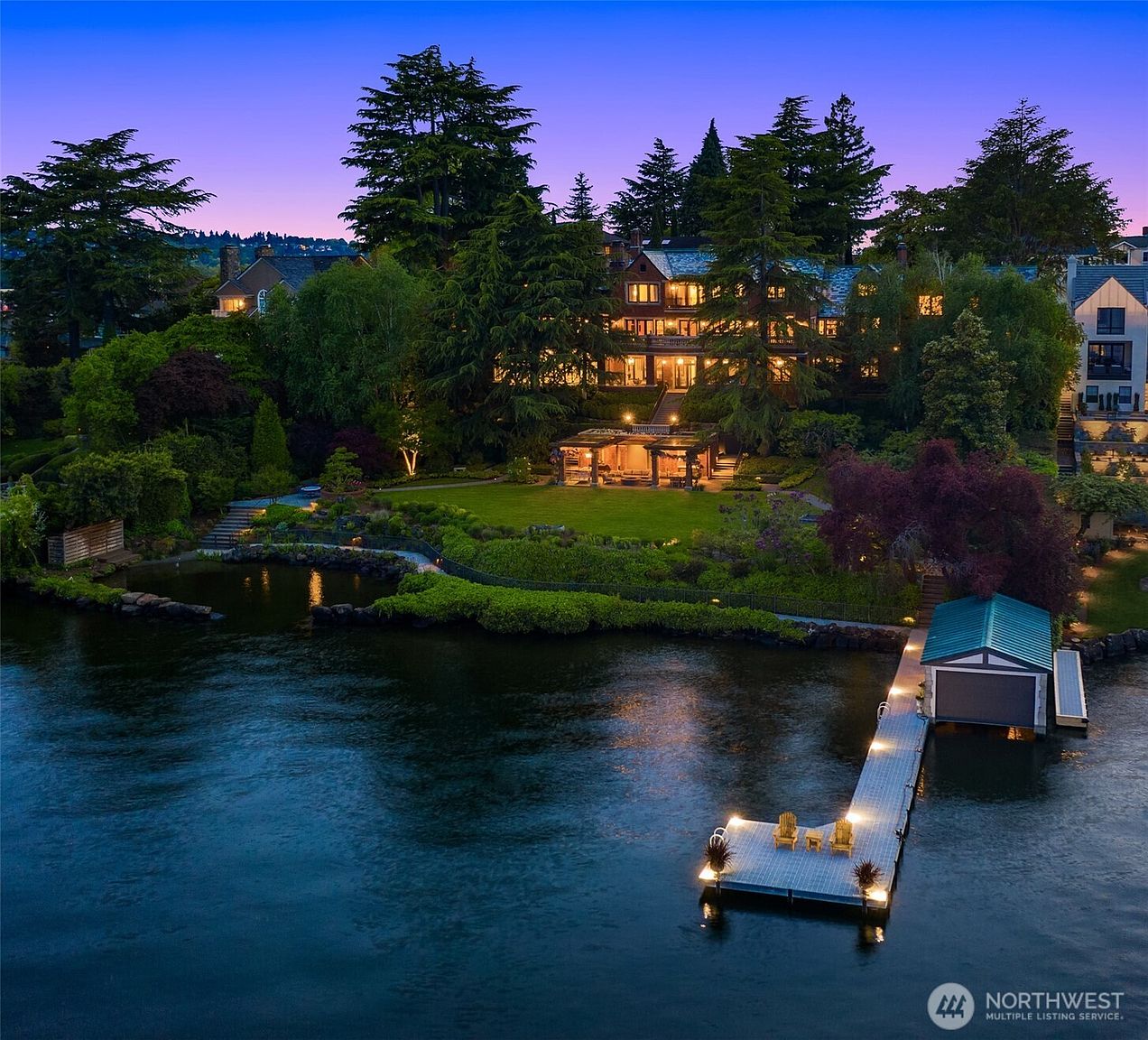 This stunning aerial view captures a luxurious waterfront estate at twilight, showcasing a grand multi-story residence nestled among lush, mature trees. The property features a meticulously landscaped lawn, a charming pergola, and a private dock extending into the calm water, complete with a boat house. The warm, inviting glow from the house and landscape lighting creates a serene and sophisticated atmosphere, highlighting the home's architectural elegance and prime lakeside location.