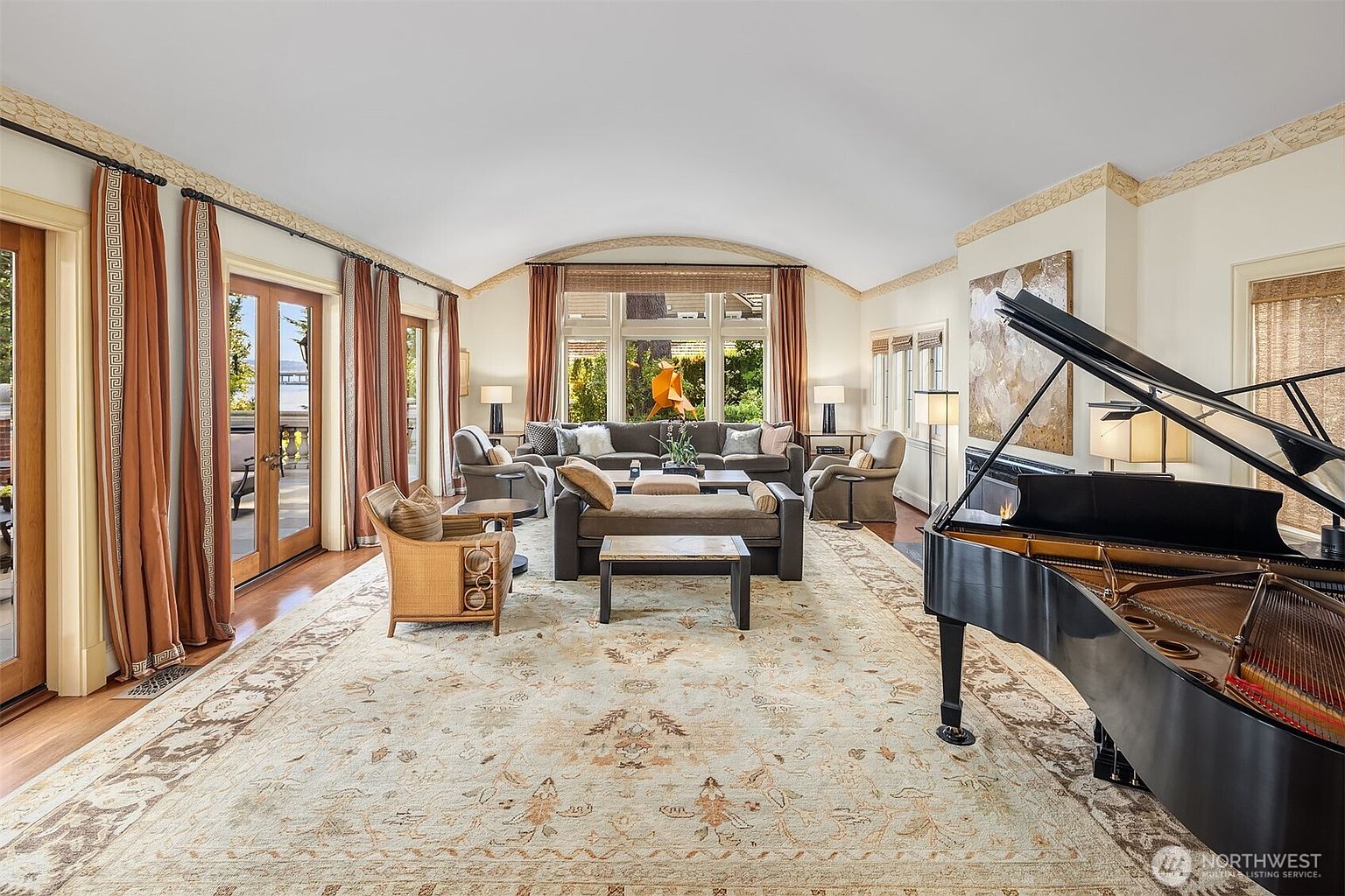 This elegant and spacious living room features a vaulted ceiling, a grand piano, and a large, ornate area rug that anchors the seating area. The room is bathed in natural light from floor-to-ceiling windows and glass doors that offer views of the outdoors, complemented by warm, neutral tones and sophisticated furnishings. The overall atmosphere is one of refined luxury and comfort, perfect for both relaxation and formal entertaining.