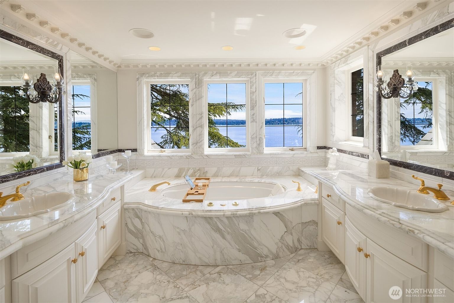 This luxurious primary bathroom features an elegant, symmetrical design centered around a large soaking tub framed by floor-to-ceiling marble and three windows overlooking a serene lake. The space is anchored by dual marble-topped vanities with gold fixtures and ornate wall sconces, reflecting a sophisticated, high-end aesthetic. The bright, airy atmosphere is enhanced by the extensive use of white marble, creating a spa-like retreat with a stunning waterfront view.
