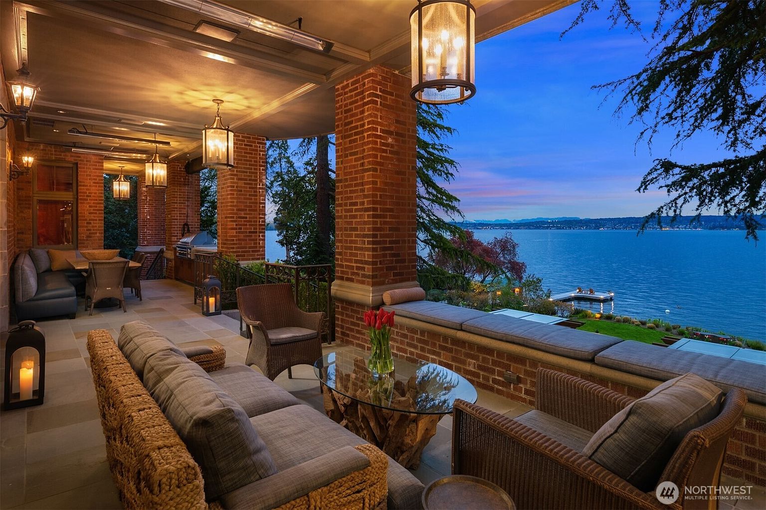 This luxurious covered patio offers a sophisticated outdoor living space with brick columns, stone flooring, and comfortable wicker furniture. The area features a built-in brick bench with plush cushions, a unique driftwood-base coffee table, and elegant hanging lanterns, all overlooking a stunning waterfront view at dusk. The atmosphere is serene and upscale, perfect for evening relaxation or entertaining.