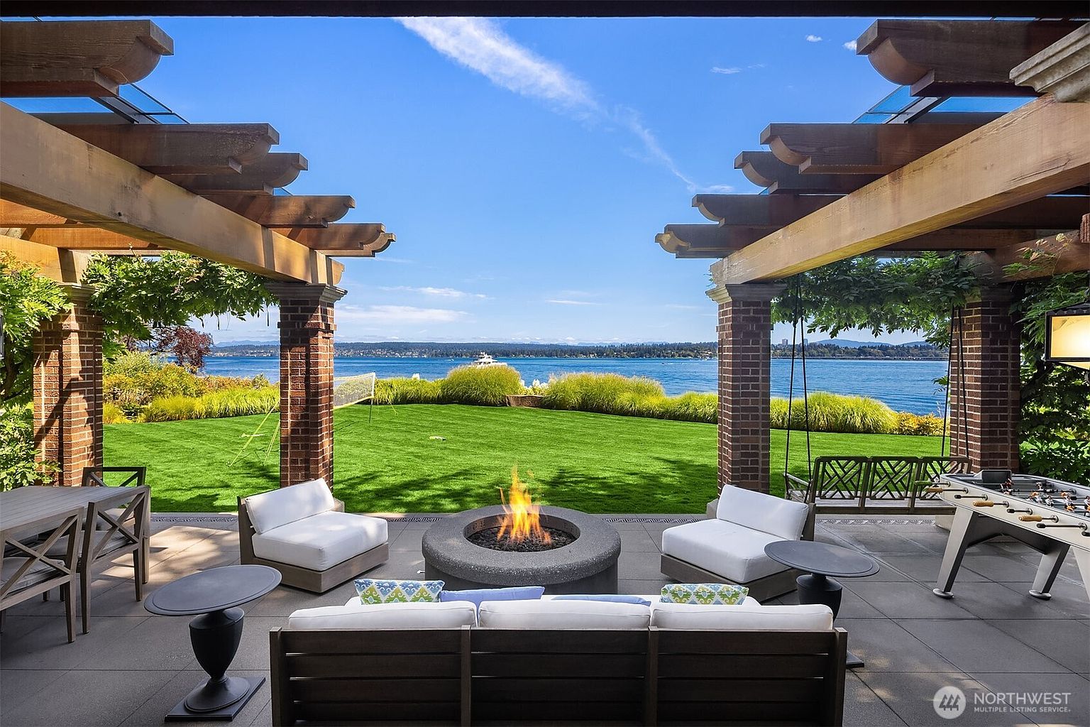 This luxurious covered patio offers a stunning, unobstructed view of a serene lake and lush green lawn, framed by elegant brick columns and a wooden pergola. The space is furnished with comfortable white lounge seating, a central circular fire pit, and a foosball table, creating an ideal setting for outdoor relaxation and entertainment. The perspective is from under the shaded structure, looking out toward the water, emphasizing the seamless transition between indoor comfort and the natural landscape.
