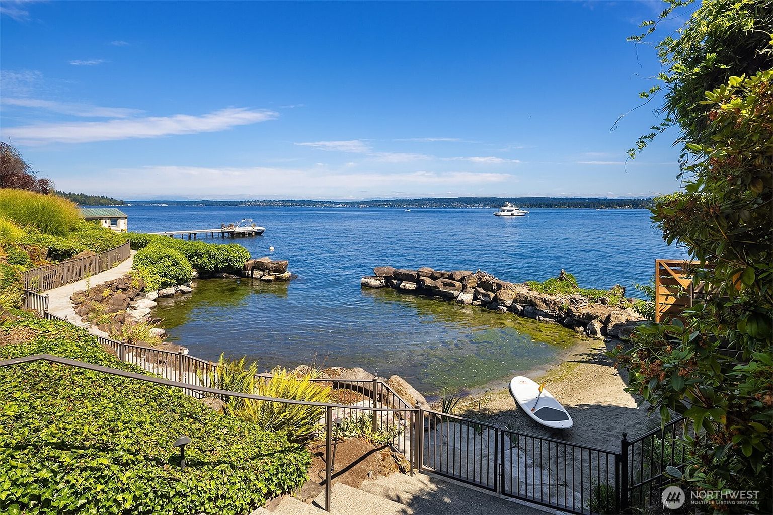 This scenic waterfront property features a private sandy beach cove framed by natural rock formations and lush landscaping. A metal gate and pathway lead down to the water, where a small boat dock and a paddleboard are visible, offering a serene and luxurious lakeside lifestyle. The perspective is elevated, looking out over the expansive blue water toward a distant shoreline under a clear, bright sky.