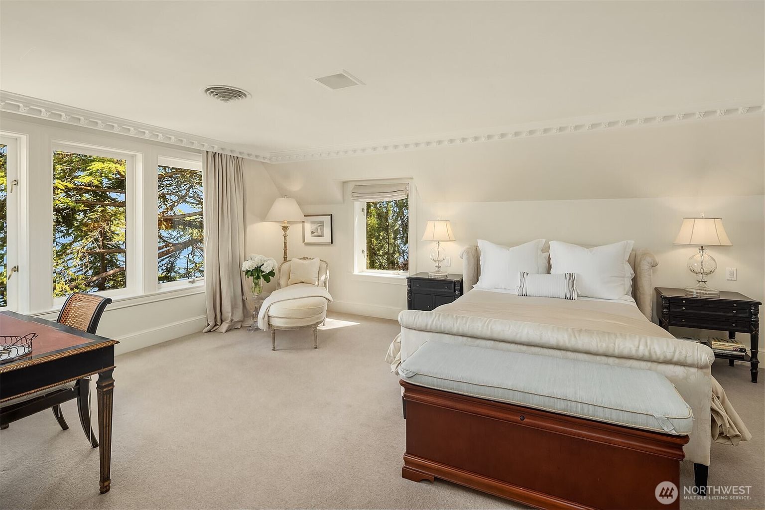 This elegant and spacious primary bedroom features a neutral color palette, plush carpeting, and a large bed with a wooden bench at its foot. The room is bathed in natural light from multiple windows that offer serene views of the surrounding trees and water, while a cozy chaise lounge provides a perfect reading nook. The sophisticated decor, including matching bedside lamps and a classic desk, creates a tranquil and luxurious atmosphere.