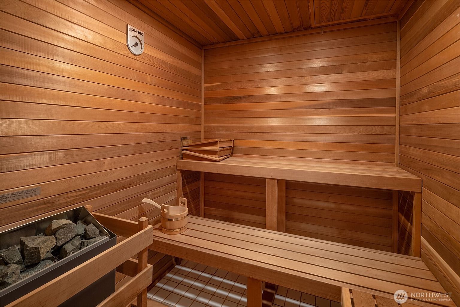 This image showcases a traditional cedar-lined sauna, featuring tiered wooden benches and a dedicated heater filled with sauna stones. A small wooden bucket and ladle sit on the lower bench, emphasizing the authentic spa-like experience. The warm, natural wood tones create a cozy and relaxing atmosphere, perfect for a home wellness retreat.