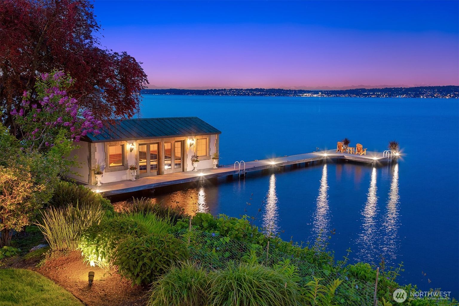 This stunning waterfront property features a charming boathouse structure connected to an expansive, illuminated wooden dock extending into a calm lake at twilight. The scene is framed by lush, manicured landscaping and vibrant purple flowering trees, creating a serene and luxurious atmosphere. The perspective is elevated, capturing the reflection of the dock lights on the water against a beautiful gradient sunset sky.