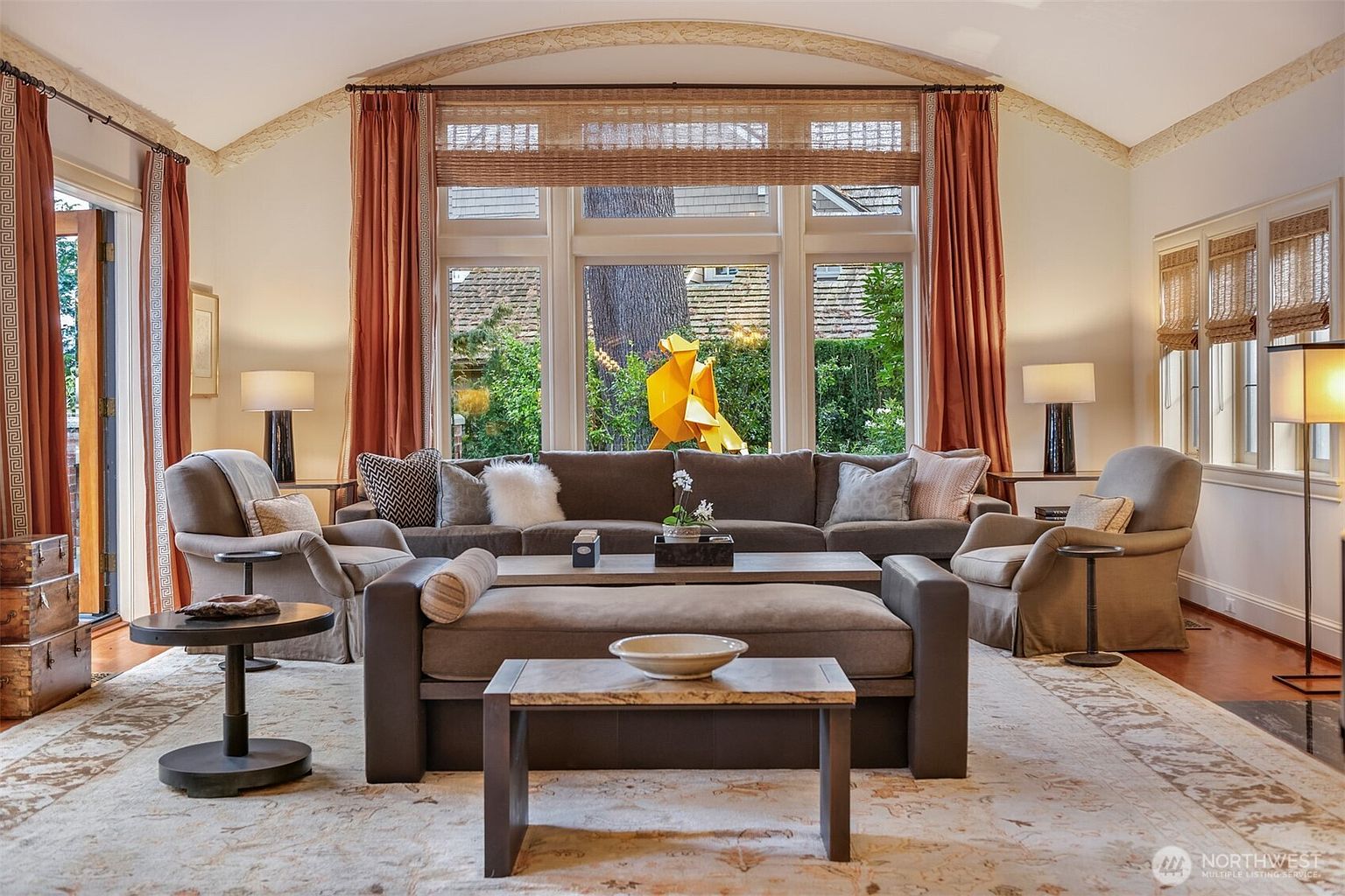 This elegant living room features a high, vaulted ceiling with decorative molding and a large, central window arrangement that floods the space with natural light. The room is anchored by a plush, neutral-toned sectional sofa and matching armchairs, complemented by a long ottoman and a low-profile coffee table on a patterned area rug. Warm, rust-colored curtains frame the windows, creating a sophisticated and inviting atmosphere perfect for relaxation or entertaining.