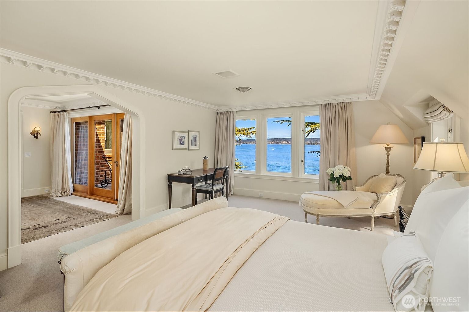 This elegant primary bedroom features a neutral color palette, plush carpeting, and a sophisticated chaise lounge positioned near large windows that offer a stunning, unobstructed view of the water. An arched entryway leads to a secondary space with glass doors, while crown molding and classic furniture pieces enhance the room's luxurious and serene atmosphere.