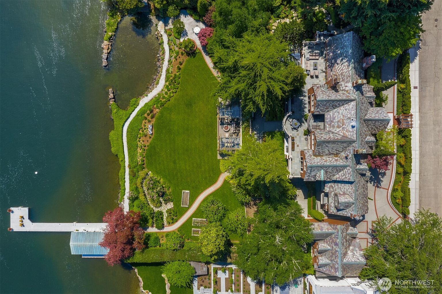 This stunning aerial perspective captures a luxurious waterfront estate, showcasing a sprawling manicured lawn, lush mature landscaping, and a private dock extending into the calm water. The grand residence features a complex, multi-gabled roofline with intricate architectural detailing, surrounded by stone pathways and elegant outdoor living spaces. The composition highlights the seamless integration between the sophisticated home and its serene, natural lakeside setting.