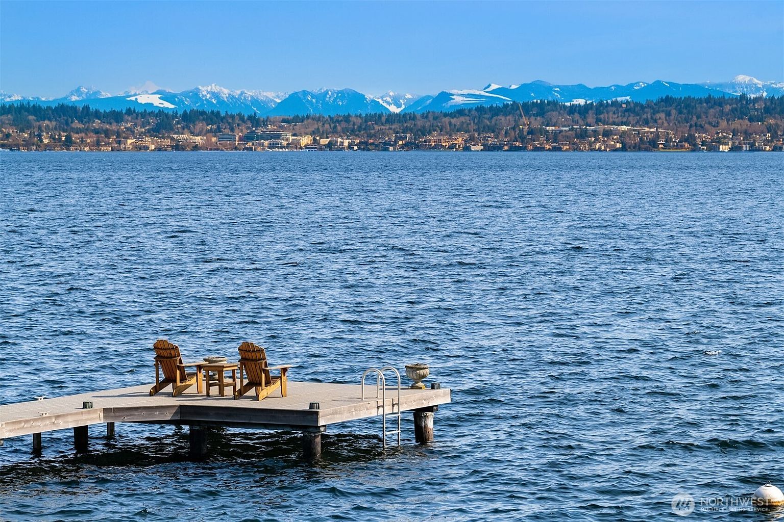 This image captures a serene, elevated view of a wooden dock extending into a vast, deep blue lake. Two Adirondack chairs and a small side table are positioned on the dock, creating a peaceful seating area that overlooks the water toward a distant, snow-capped mountain range and a shoreline community. The perspective is wide and cinematic, emphasizing the tranquil waterfront lifestyle and the expansive natural scenery.